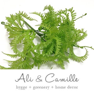 May include: A bunch of green artificial fern sprigs. The ferns are arranged in a loose, natural-looking cluster. The ferns are a light green color and have a delicate, feathery texture.