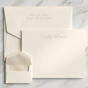 May include: A set of cream-colored stationery featuring a flat note card embossed with the name Emily Woods and a matching envelope embossed with the address 520 Circle Drive, Tampa, Florida 33629, alongside a smaller, empty cream-colored envelope.