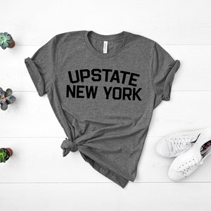 May include: A heather gray t-shirt with the words "UPSTATE NEW YORK" printed in black. The shirt is knotted at the waist and paired with white sneakers. Small potted succulents are arranged on a white wooden surface.