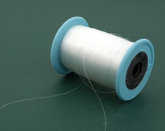May include: A spool of clear, thin nylon monofilament thread wound around a light blue plastic core with a black center. The thread extends from the spool in a loose, curved loop, showcasing the transparent and flexible nature of the material.