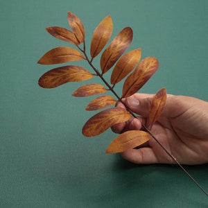 Puede incluir: Una mano sosteniendo una rama de hojas marrones y naranjas sobre un fondo verde.
