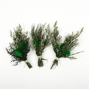 May include: Three small bouquets of dried greenery, including sprigs of green and tan grasses, and dark green accents. Each bundle is tied with twine. The background is plain white.