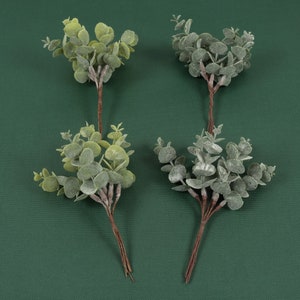 May include: Four sprigs of artificial eucalyptus greenery with a frosted finish. The sprigs are arranged on a green background.