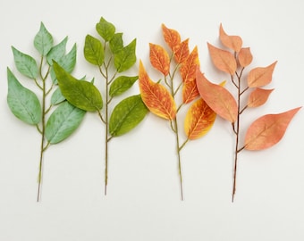 May include: Four artificial leaf stems in varying colors: light green, green, orange, and peach. The leaves are arranged on thin brown stems against a white background. The leaves' colors transition from spring to autumn.