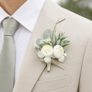 Op de afbeelding: Een crèmekleurige corsage met twee witte rozen, groen en een lint is vastgemaakt aan een beige jasje. Een lichtgroene stropdas en een wit overhemd zijn ook zichtbaar.