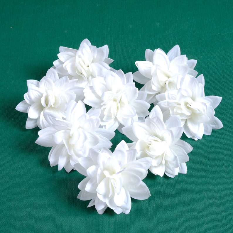 Puede incluir: Un grupo de siete flores artificiales blancas dispuestas en forma de coraz&oacute;n sobre un fondo verde.