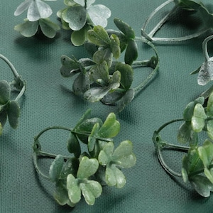 May include: A close-up of several green artificial plant sprigs with wire stems. The sprigs are arranged on a green textured surface.