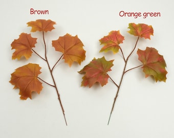 May include: Two artificial leaf branches. One branch features brown leaves, and the other has orange and green leaves. The leaves have a maple leaf shape. The branches are brown and green, and the leaves are various shades of orange, brown, and green.