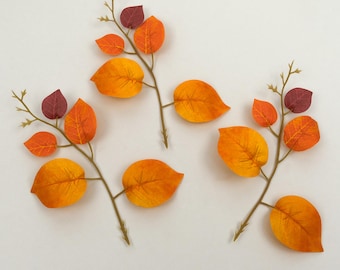 May include: Three artificial leaf branches with orange, yellow, and burgundy leaves. The leaves are arranged on thin, brown stems, creating a fall-themed decorative element. The leaves have a gradient of colors, adding depth and visual interest.