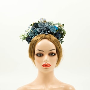Op de afbeelding: Een bloemenkrans met een assortiment van blauwe en groene kunstbloemen en bladeren. De krans bestaat uit grote, getextureerde blauwe bloemen, kleinere blauwe en witte bloesems en groene bladeren. De krans wordt gedragen op een mannequinhoofd met lichtbruin haar.