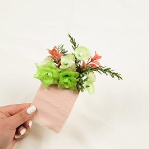 May include: A floral boutonniere featuring light green artificial roses, small white buds, and coral-colored foliage accents. The stems are wrapped in a wide, pale pink fabric ribbon with a textured, frayed edge finish for a soft appearance.