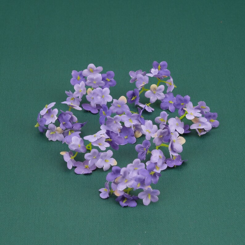 May include: A cluster of purple and white artificial flowers arranged in a star shape on a green background.