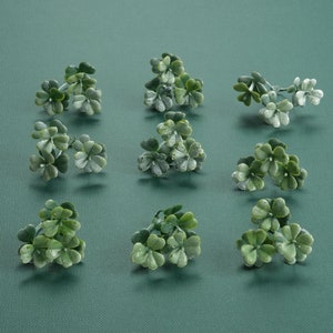 May include: Twelve small green artificial shamrock sprigs on a green background.
