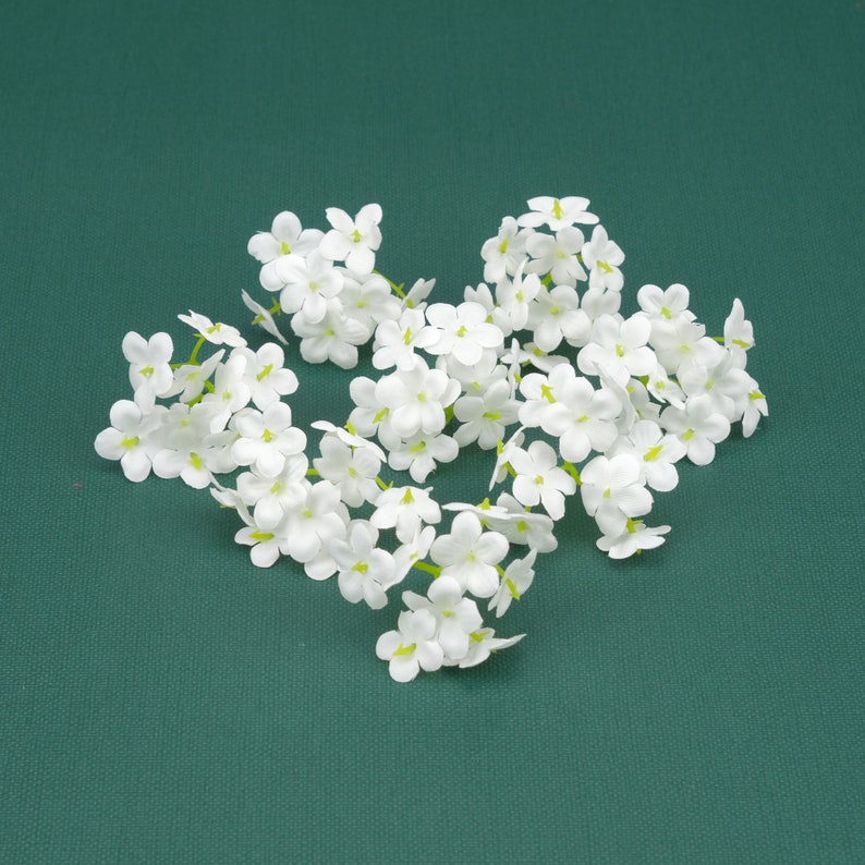 May include: A cluster of white artificial flowers with green stems, arranged in a heart shape on a green background.
