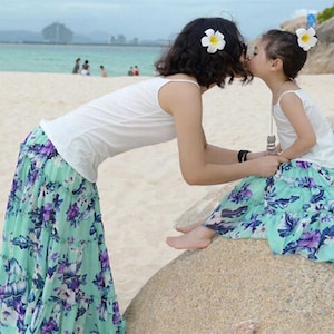 May include: A mother and daughter wearing matching floral print skirts and white tank tops. They are both wearing white flower hair clips. The mother is kissing the daughter on the cheek.