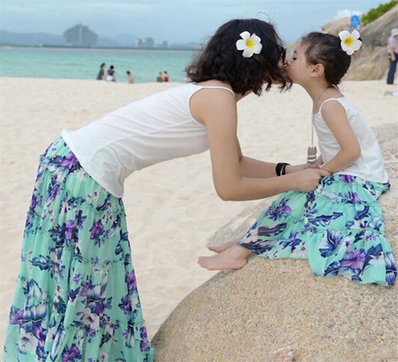 May include: A mother and daughter wearing matching floral print skirts and white tank tops. They are both wearing white flower hair clips. The mother is kissing the daughter on the cheek.