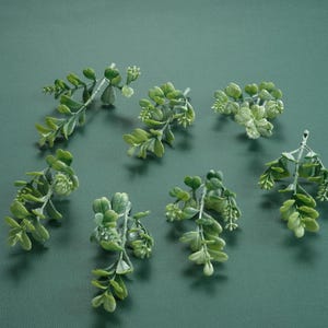 May include: A collection of seven sprigs of green artificial greenery, arranged on a green background.