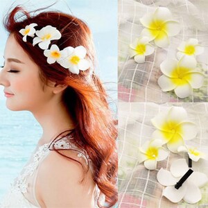 May include: White flower hair clips with yellow centers. The clips are arranged in a row on a woman's hair. The woman is wearing a white dress.