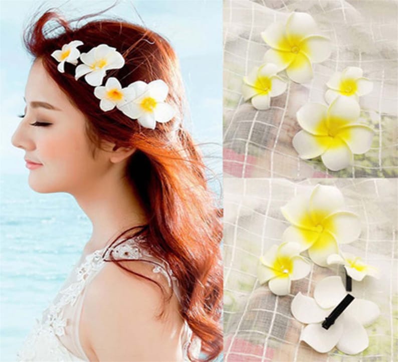 May include: White flower hair clips with yellow centers. The clips are arranged in a row on a woman's hair. The woman is wearing a white dress.