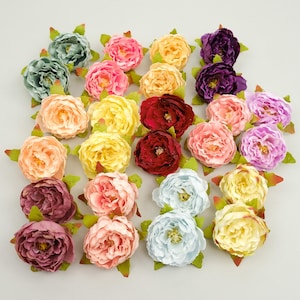 May include: A collection of artificial peony flower heads in various colors, including pink, yellow, red, purple, and blue. Each flower has green leaves at the base. The flowers are arranged on a light-colored surface.