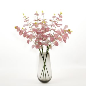Puede incluir: Un jarrón de cristal transparente contiene un ramo de ramas de eucalipto artificiales. Las hojas son de un tono rosa suave, con pequeños capullos amarillos en las puntas. El jarrón tiene un tinte gris ahumado y forma de lágrima.