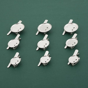 May include: Ten silver-toned metal blank round pin backings with a smooth surface. The pin backings are arranged in three rows on a green surface.