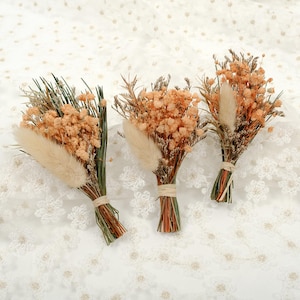 Op de afbeelding: Drie kleine boeketten van gedroogde bloemen. Elk boeket heeft perzikroze gipskruid, gedroogde grassen en een pluizig, beige accent. De stelen zijn gebonden met touw. De boeketten zijn gerangschikt op een wit oppervlak met kantpatroon.