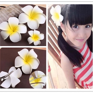 May include: Four white and yellow plumeria flower hair clips. The flowers are made of fabric and have a realistic appearance. The clips are attached to the back of the flowers.