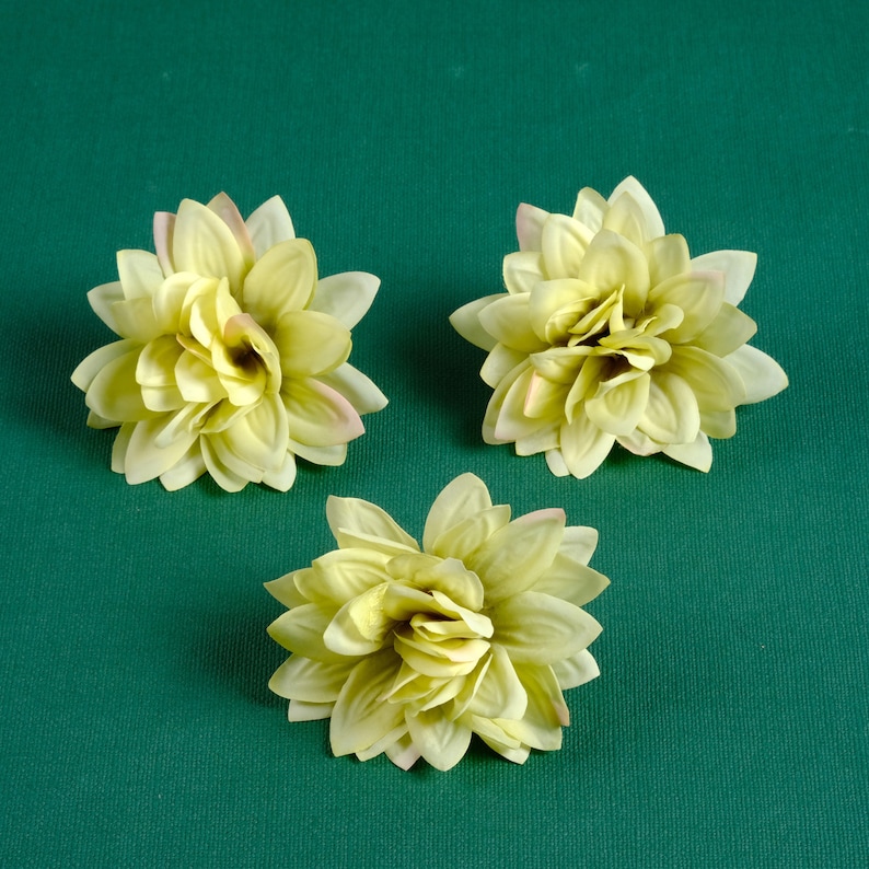 Puede incluir: Tres flores artificiales de color amarillo p&aacute;lido con un ligero tono rosado en los bordes de los p&eacute;talos. Las flores est&aacute;n dispuestas sobre un fondo verde.