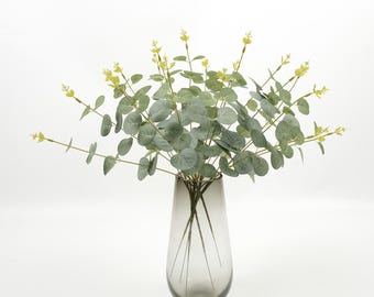 Op de afbeelding: Een boeket van kunstmatige eucalyptus in een grijze glazen vaas. De eucalyptus heeft ronde, grijsgroene bladeren en kleine gele knoppen. De vaas is druppelvormig en staat op een witte achtergrond. De opstelling is een decoratief woonaccessoire.