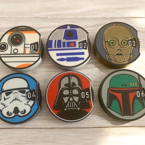 Könnte beinhalten: Sechs runde Flaschenverschlüsse mit Star Wars-Motiven. Jeder Verschluss zeigt einen anderen Charakter, darunter BB-8, R2-D2, C-3PO, einen Sturmtruppler, Darth Vader und Boba Fett. Jeder Verschluss ist mit einer Nummer von 01 bis 06 versehen.