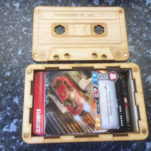 May include: A wooden cassette tape-shaped box, open to reveal trading cards. The box is light brown with etched details, including the text "TRANSFORMERS MIX TAPE." The cards feature a red car and character details. The box is designed to hold and display collectible cards.