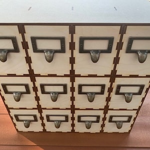 May include: A wooden box with twelve small drawers, each with a metal label holder and a pull handle. The drawers are arranged in a 3x4 grid.