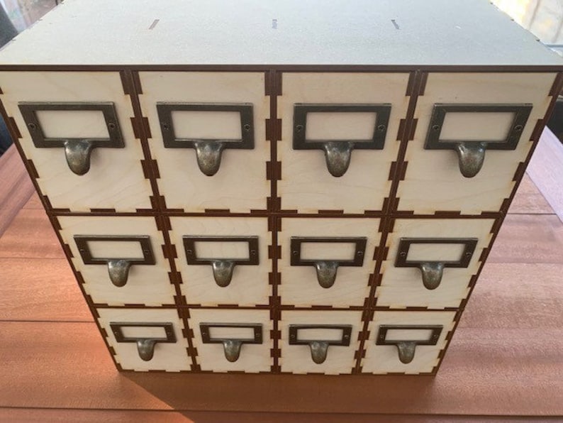 May include: A wooden box with twelve small drawers, each with a metal label holder and a pull handle. The drawers are arranged in a 3x4 grid.