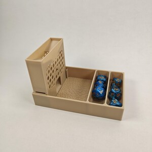 以下が含まれることがあります： ベージュのプラスチック製のサイコロタワーとトレイ。トレイにはサイコロを入れるための仕切りがあります。タワーはハニカムデザインで、トレイはテクスチャ加工されています。仕切りには、青と金のサイコロがいくつか入っています。