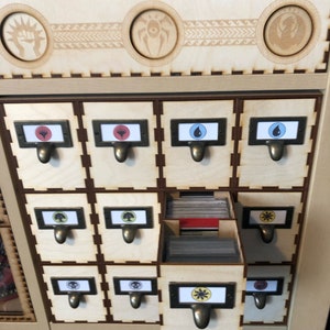 May include: A wooden box with 12 drawers for storing trading cards. Each drawer has a different symbol on the front, including a fist, a spider, a dragon, a drop of water, a tree, a skull, a sun, and a heart. The drawers are labeled with the symbols.