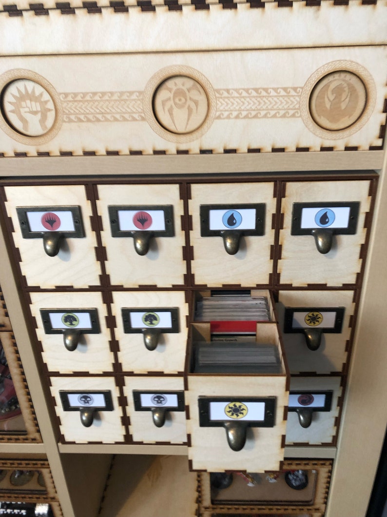 May include: A wooden box with 12 drawers for storing trading cards. Each drawer has a different symbol on the front, including a fist, a spider, a dragon, a drop of water, a tree, a skull, a sun, and a heart. The drawers are labeled with the symbols.