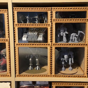 Könnte beinhalten: Ein Holzdisplay mit mehreren Fächern, die jeweils ein Miniaturmodell eines Roboters oder Fahrzeugs enthalten. Die Modelle sind in verschiedenen Grautönen und Schwarz lackiert.