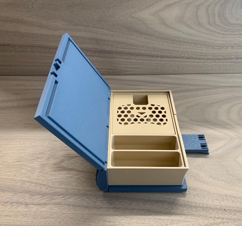 以下が含まれることがあります： 蓋にハニカム模様が施された青とベージュの収納ボックス。ボックスには2つのコンパートメントとヒンジ付きの蓋があります。