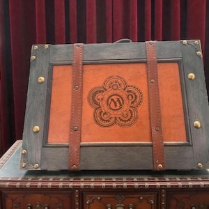 May include: A rectangular wooden chest featuring a dark gray frame with metallic corner accents and two vertical brown leather straps. The center panel is light brown with a black circular emblem containing the letter M and intricate gear designs.
