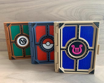 May include: Three closed books with different designs. The book on the left has a green cover with a white symbol. The middle book has a red cover with a white and black symbol. The book on the right has a blue cover with a white and pink symbol.