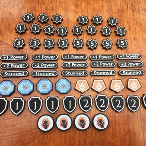 Puede incluir: Una colección de fichas de juego blancas y negras con varios números y símbolos. Las fichas incluyen los números 1, 2, 3 y 5, así como símbolos como "+1 Power", "+2 Power" y "Stunned".
