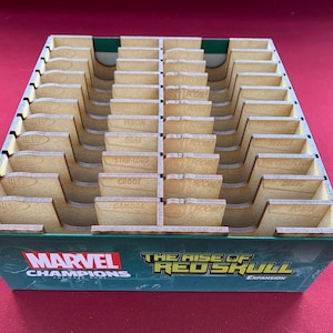 Puede incluir: Un organizador de madera para el juego de mesa Marvel Champions: The Rise of Red Skull. La caja verde presenta el logotipo de Marvel Champions y el título de la expansión. El organizador tiene múltiples compartimentos con nombres de personajes.