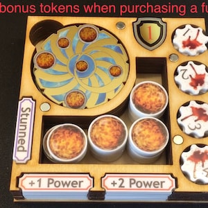 May include: Wooden organizer for a board game with compartments for different tokens. The organizer has a compartment for "Stunned" tokens, a compartment for "Power" tokens, and a compartment for "Blood" tokens. The organizer also has a compartment for a spinning wheel with a gold and blue design.