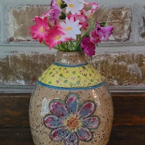 May include: A ceramic vase with a floral design and a yellow dotted top, holding a bouquet of pink, white, and purple flowers. The vase has a hand-painted flower on the front and a blue stripe.
