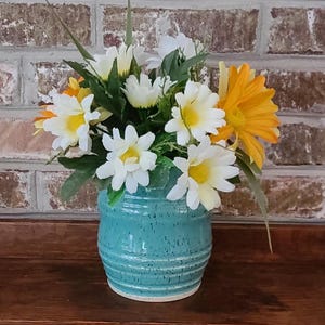 May include: A turquoise ceramic vase holds a vibrant arrangement of artificial flowers. The bouquet features white daisies with yellow centers, and bright yellow gerbera daisies. The vase has a speckled finish and horizontal grooves.