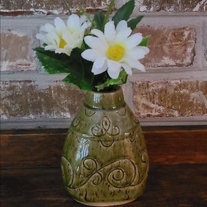 May include: A small, green ceramic vase with embossed floral designs, holding white and yellow artificial daisies with green leaves. The vase is on a wooden surface, with a brick wall in the background.