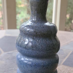 May include: A handcrafted ceramic vase with a unique, rounded shape. The vase features a speckled blue glaze with darker blue and gray accents. The small vase is suitable for displaying small flowers or as a decorative accent piece.