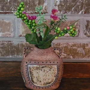 May include: A small, speckled, ceramic vase with two handles, holding artificial flowers. The flowers include pink blooms, green berries, and green leaves. The vase has a light brown color with a lighter, rectangular design on the front.