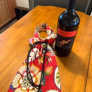 Op de afbeelding: Een rode trekzak met een bloemenpatroon en zwarte trekkoorden. De zak ligt op een houten tafel naast een fles "Yellow Tail" Cabernet Sauvignon wijn.